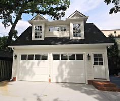 a white garage with two windows on the top floor and one story above it, in front of a large tree