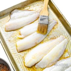 some fish are sitting on a baking pan with a brush in one hand and seasonings in the other