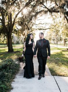 a man and woman holding hands walking down a sidewalk in front of trees with spanish moss