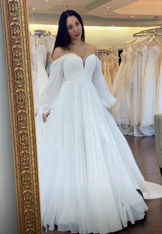 a woman standing in front of a mirror wearing a white dress with long sleeves and pleated skirt