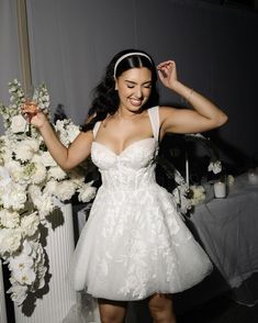 a woman in a short white dress standing next to a table with flowers on it