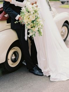 a bride and groom standing next to an old car with flowers on the front, dressed in black tuxedo