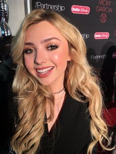 a woman with long blonde hair and black shirt smiling at the camera while standing in front of a red carpet