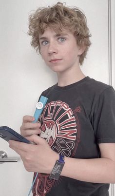 a young boy holding an electronic device and looking at the camera while standing in front of a white wall