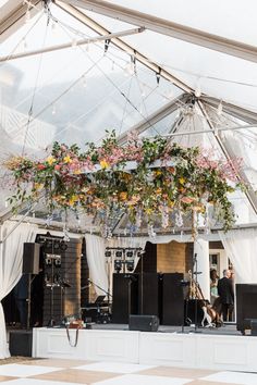 a white tent with flowers hanging from it's ceiling and two people standing on the stage