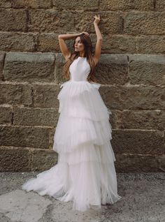 a woman in a white dress leaning against a stone wall with her arms up and hands behind her head