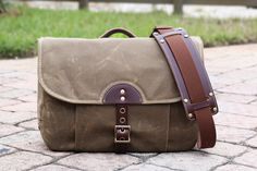 a brown and tan messenger bag sitting on top of a brick floor next to grass