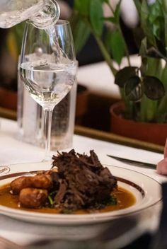 a white plate topped with meat and gravy next to a glass of wine