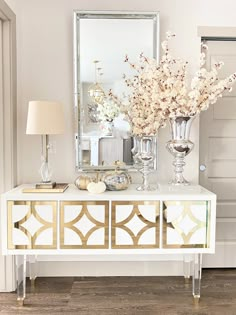 a white and gold sideboard with flowers on it in front of a large mirror