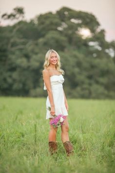 a woman standing in the middle of a field with her boots on and flowers in her hand