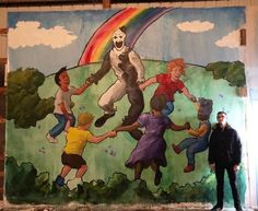 a man standing in front of a painting of children playing with a bear and rainbow