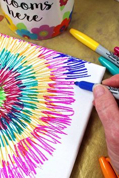 a person is drawing on a canvas with crayons and marker pens in front of a coffee mug