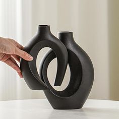 two black vases sitting on top of a table next to each other, one being held by a person's hand