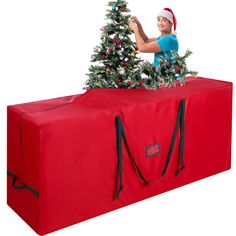 a woman is decorating a christmas tree on top of a large red bag with black straps