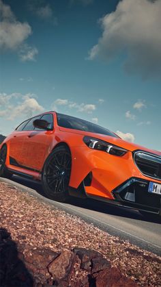 an orange car driving down a road next to some rocks and grass with clouds in the background