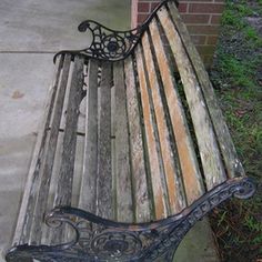 an old wooden bench with wrought iron arm rests on the sidewalk near some grass and bushes