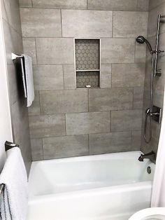 a white toilet sitting next to a bath tub under a window in a bathroom with gray tiles on the walls