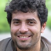 a man with curly hair smiling at the camera while wearing a gray shirt and jacket