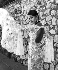 a woman standing next to a stone wall holding onto some white lace on her dress