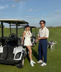 a man and woman standing next to a golf cart on a green field with a dog