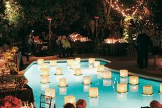 people sitting at tables in front of a pool with lit candles on the water and around them