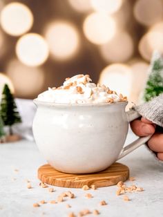 a person holding a mug with whipped cream in it and christmas decorations around the cup