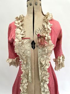 "This rare and absolutely lovely antique late Victorian over dress is made from pink figured silk with cream figured silk highlights at the bodice. The edges of the pink silk fabric are trimmed with delightful ruffled cream coloured silk and the sleeve ends are trimmed with ruffled silk and lace. There is a beautiful flower corsage to the left hand side of the bodice. The inside of the bodice is boned with what looks to be steel and is lined with cotton. The design of the dress has swags of fabric to each side which falls beautifully when it is displayed on a mannequin/dress form, as does the back of the dress which has a full-length central pleat. This would be a lovely addition to a collection of antique clothing.  It has an interior label for the department store George Henry Lee and Co Pink Victorian Rococo Dress For Costume, Pink Rococo Victorian Dress For Costume, Pink Rococo Victorian Dress Costume, Pink Fitted Rococo Victorian Dress, Pink Victorian Marie Antoinette Costume Dress, Pink Rococo Victorian Dress, Pink Victorian Dress With Historical Design For Wedding, Vintage Pink Costume Dresses, Elegant Pink Victorian Dress With Historical Design