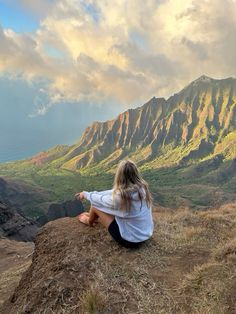 a woman sitting on top of a hill with mountains in the background and clouds overhead