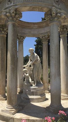 a statue in the middle of some pillars with pink flowers around it and blue sky above