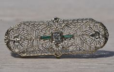 The El Granada: Antique Filigree Brooch with Old Cut Diamond and Emeralds. The pin features an old mine cut central diamond in a square box finished with milgrain set among an intricate filigree pattern complimented by foliage and floral motif. Elongated French cut emeralds finish center. The brooch is crafted in 14 karat white gold and measures 1 3/4 inches by 1/2 inch. Love this piece, but don't have the money to spend right now? We offer FREE layaway on every item in our shop. With just 20% d Heirloom Filigree Brooches For Formal Occasions, Heirloom Filigree Brooches For Anniversary, Art Deco Green Brooches For Wedding, Elegant Green Filigree Brooches, Heirloom Engraved Brooch For Wedding, Heirloom Wedding Brooches With Intricate Design, Heirloom Engraved Brooches For Wedding, Green Art Deco Brooches For Wedding, Vintage Diamond Brooches With Filigree