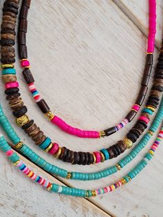 three necklaces with different colored beads are on a white wooden surface and one has a pink ribbon