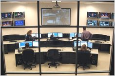 people sitting at desks in an office setting with multiple monitors and laptops on the wall