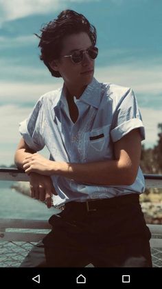 a man with his arms crossed standing in front of the ocean wearing sunglasses and a button up shirt