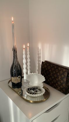 two white candles are sitting on a tray next to a bottle and purse in a room