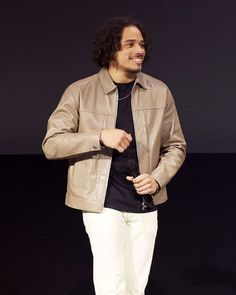 a man in a tan leather jacket and white pants standing on a stage with his hands in his pockets