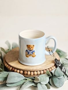 a coffee mug with a teddy bear on it sitting on a wooden coaster surrounded by greenery