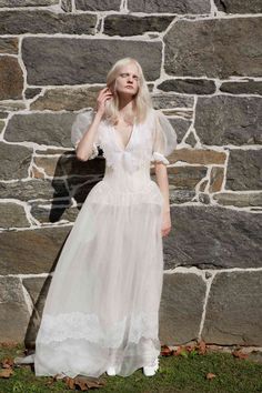 a woman standing in front of a stone wall wearing a white dress and holding her hand up to her ear