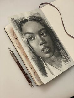 a pencil drawing of a woman's face on top of a notebook with a pen next to it