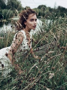 a woman sitting in tall grass next to a body of water