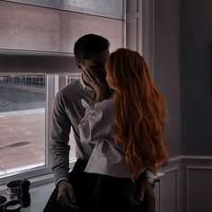 a man and woman standing next to each other in front of a window looking out the window