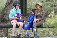 three people sitting on a bench with a purple teddy bear in their lap and one person wearing a helmet