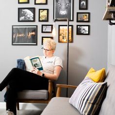 a woman sitting on a couch reading a book in front of a wall full of pictures