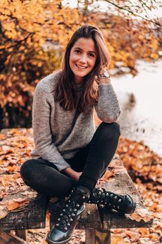 a woman sitting on top of a wooden bench in front of some trees and leaves