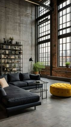 an industrial living room with large windows and black leather couches, coffee table, and yellow ottoman