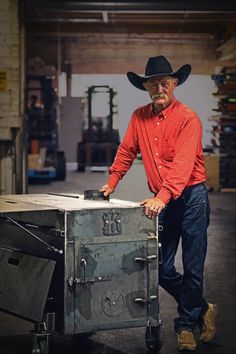 a man in a red shirt and cowboy hat standing next to a metal box with wheels