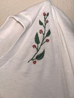 a white t - shirt with red flowers and leaves embroidered on the front, sitting against a wall