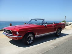 an old red mustang convertible parked on the side of the road next to the ocean