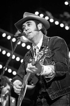 a man with a cowboy hat on singing into a microphone and playing an acoustic guitar