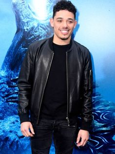 a man standing in front of a blue background wearing a black shirt and leather jacket