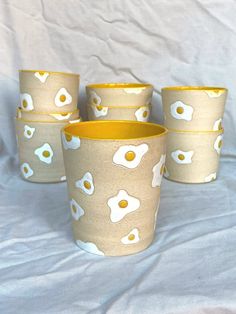 four cups with eggs on them are sitting on a white cloth covered tablecloth, one is yellow and the other has an egg pattern